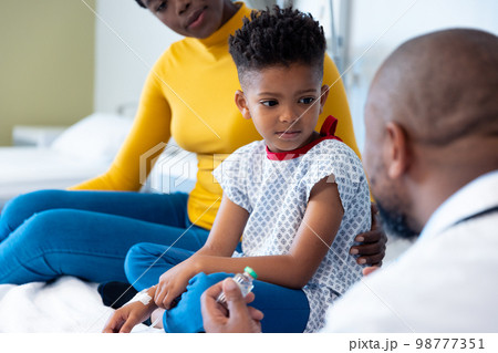 African american male doctor holding vaccine talking to boy patient, with mother 98777351