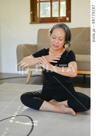 60s aged Asian woman massaging her wrist, injured hand during yoga practice at home. 98779287