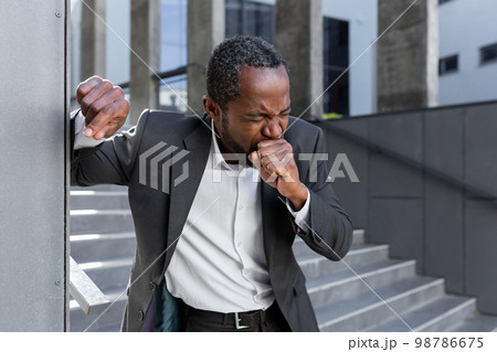 Cough attack. An African-American businessman in a suit is holding on to the office wall outside, coughing hard, covering his mouth with his hand. feels chest pain. Cough attack. An African-American businessman in a suit is holding on to the office wall outside, coughing hard, covering his mouth with his hand. feels chest pain. 98786675