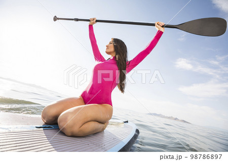 Woman in pink swimsuit kneeling on paddleboard on lake 98786897