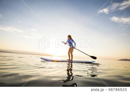 Woman in blue swimsuit paddleboarding on lake at sunset 98786920