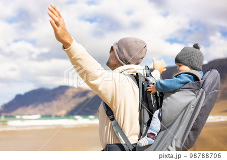 Young father rising hands to the sky while enjoying pure nature carrying his infant baby boy sun in backpack on windy sandy beach of Famara, Lanzarote island, Spain. Family travel concept 98788706