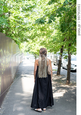 A girl in a black dress and long afro-braids walks along a summer street, rear view A girl in a black dress and long afro-braids walks along a summer street, rear view 98791081