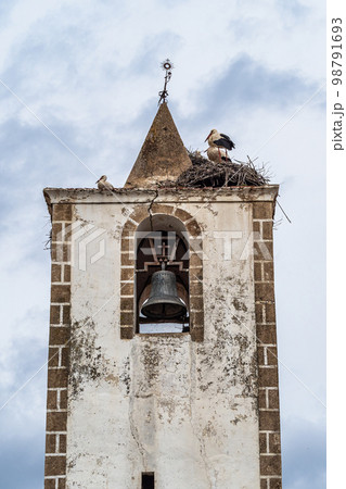 Nest of storks on top of old stone bell tower with iron cross at Herreruela, Extremadura in Spain 98791693