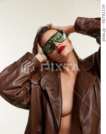 Attractive sensual portrait of beautiful young brunette girl with wet hair in stylish leather jacket, green sunglasses posing over grey studio background. Concept of beauty, fashion 98792139