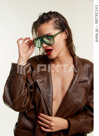 Flirty look. Beautiful young brunette girl in stylish leather jacket and green sunglasses posing over grey studio background. Concept of beauty, fashion Flirty look. Beautiful young brunette girl in stylish leather jacket and green sunglasses posing over grey studio background. Concept of beauty, fashion 98792141