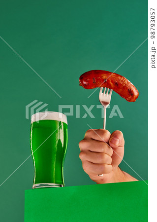 Glass with foam green beer and make hand with sausage on fork over green background. Traditional food and drink combination. Concept of st patrick's day celebration, brewery, traditions 98793057