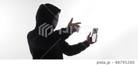 Hacker Anonymous and face mask with smartphone in hand. Man in black hood shirt holding and using mobile phone on white background. Represent cyber crime data hacking or stealing personal data concept Hacker Anonymous and face mask with smartphone in hand. Man in black hood shirt holding and using mobile phone on white background. Represent cyber crime data hacking or stealing personal data concept 98795260