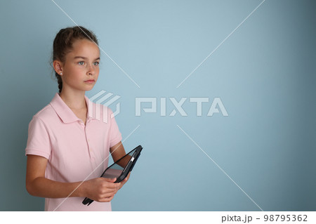Little emotional teenage girl in pink shirt 11, 12 years old on an isolated blue background with a tablet in her hands.Children's studio portrait. Place the text to copy the place for the inscription 98795362