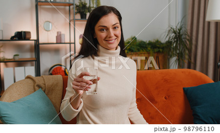 Thirsty young woman sitting at home holding glass of natural aqua make sips drinking still water Thirsty young woman sitting at home holding glass of natural aqua make sips drinking still water 98796110