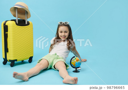 Smiling Caucasian adorable child traveler tourist adventurer, cute mischievous little girl rejoicing while going for summer getaway, isolated blue color background, near a yellow suitcase and globe 98796665