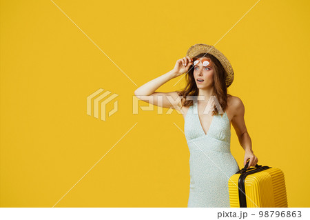 Young lady in sunglasses and summer hat holding suitcase and looking at copy space on yellow studio background 98796863