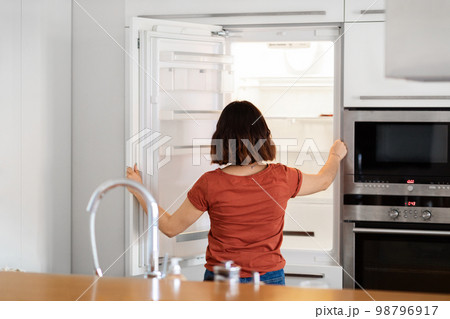 Young Woman Standing In Kitchen And Looking Inside Of Empty Fridge Young Woman Standing In Kitchen And Looking Inside Of Empty Fridge 98796917