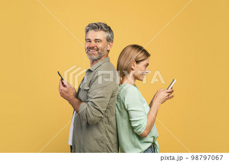 Modern lifestyle. Happy middle aged couple standing back to back, using their cellphones, man smiling at camera Modern lifestyle. Happy middle aged couple standing back to back, using their cellphones, man smiling at camera 98797067