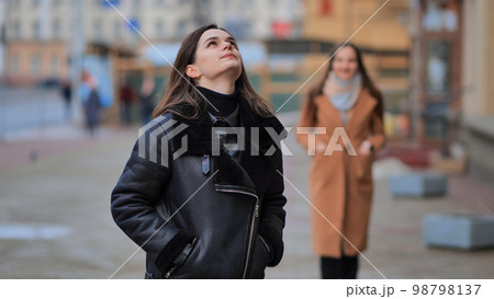 A friend comes up from behind her waiting girlfriend on a city street. 98798137