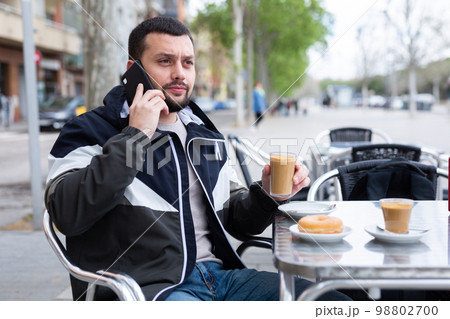 Young man talking on phone during breakfast in street cafe Young man talking on phone during breakfast in street cafe 98802700