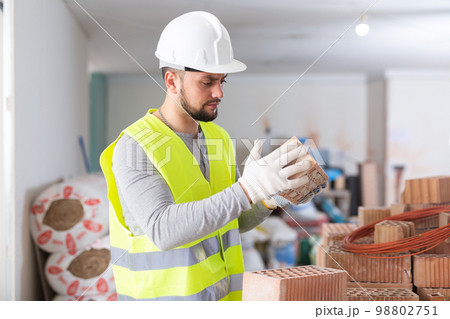 Builder in blue workwear planning erection of partition wall in building under reconstruction, preparing red bricks for work 98802751