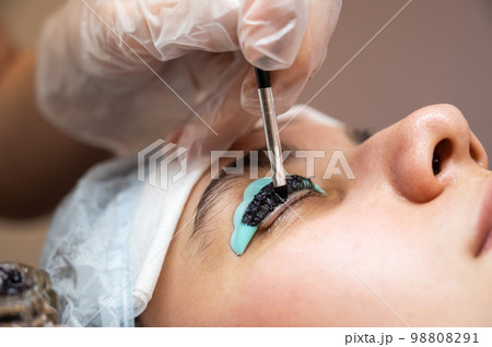 Close-up portrait of a woman on eyelash lamination procedure. The master applies tint to the eyelashes.  98808291