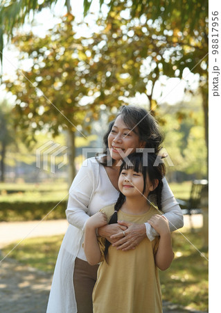 Happy middle age grandmother embracing grandchild while walking in public park surrounded by green trees at sunlight morning Happy middle age grandmother embracing grandchild while walking in public park surrounded by green trees at sunlight morning 98817956