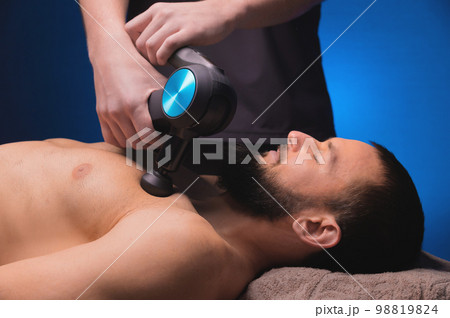 Athletic young man lies on his back, his chest is massaged with a massage gun in the salon, he warms up after training, close-up Athletic young man lies on his back, his chest is massaged with a massage gun in the salon, he warms up after training, close-up 98819824