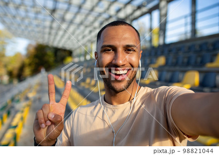 Sportsman runner in stadium taking selfie photo and talking on video call with friends, hispanic man looking at camera and smiling recording sports blog, young man after active training. 98821044