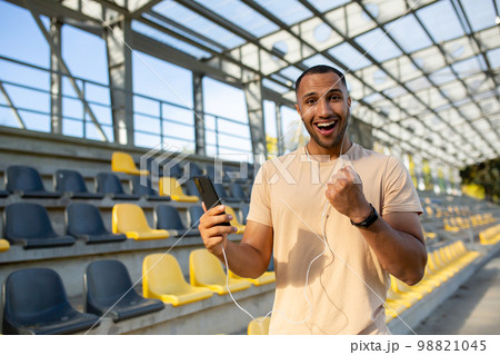 A successful man, satisfied with the result of training, looks at the camera and smiles, Hispanic holds a phone in hands uses an online sports program, an athlete holds his hand up in achievement. A successful man, satisfied with the result of training, looks at the camera and smiles, Hispanic holds a phone in hands uses an online sports program, an athlete holds his hand up in achievement. 98821045