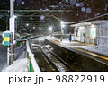 雪降る夜の無人駅に佇む帽子をかぶり青いコートを着た男性 98822919