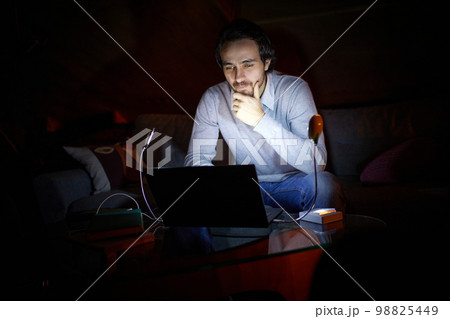 Adjusting to unstable electricity. Man, freelancer sitting on sofa at home in evening and working on laptop with battery lamp light. Blackout, no electricity Adjusting to unstable electricity. Man, freelancer sitting on sofa at home in evening and working on laptop with battery lamp light. Blackout, no electricity 98825449