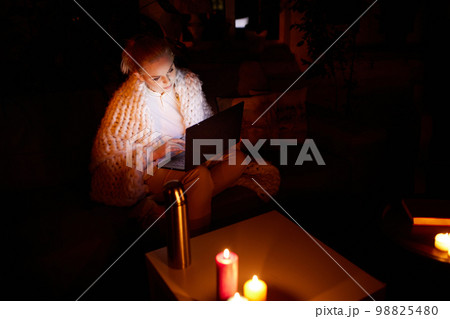 Cozy evening without electricity. Young beautiful girl sitting on sofa in warm plaid at home in the evening and working on laptop with candle light. Blackout. 98825480