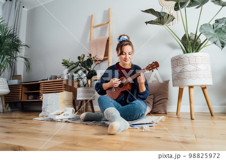 Young Woman using mobile phone while playing ukulele guitar in modern Scandi interior home. Online learning concept. Tutorial lessons via mobile application. New Hobby, resolution to learn new skills. 98825972