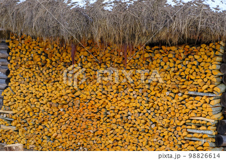 A large amount of split firewood piled up in a large neat stack along the wall of a thatched house 98826614