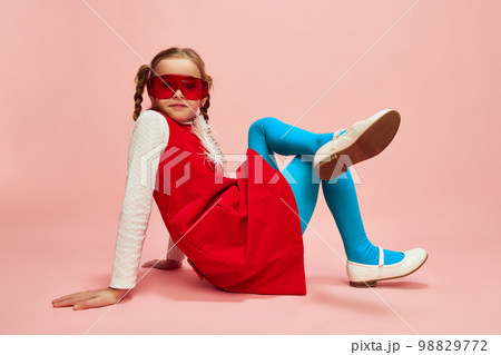 Bright style. Beautiful little girl, child in red dress, blue tights and red glasses posing over pink studio background. Concept of emotions, childhood 98829772