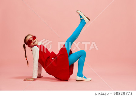 Playful, beautiful kid. Girl, child in red dress, blue tights and red glasses posing over pink studio background. Concept of emotions, childhood 98829773