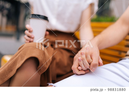Close-up cropped shot of unrecognizable happy young couple in love holding hands sitting on bench and drinking takeaway coffee enjoying time together outdoors on summer day, blurred background. 98831880