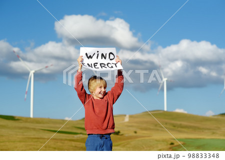 Eco activist boy with banner "Wind Energy" on background of power stations for renewable electric energy production. Child and windmills. Wind turbines for generation electricity. Green energy 98833348