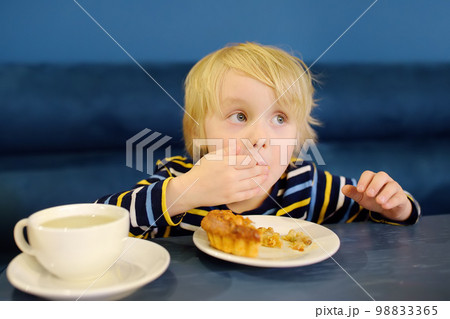 Cute blonde boy eating pie and drinking tea in cafe or restaurant. Child eating with his hands. Healthy meal for kids. 98833365