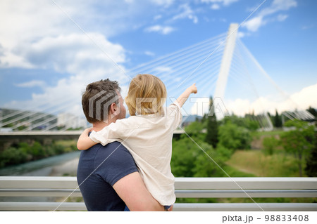 Little child and middle age father are admiring of cable stayed bridge. Boy is pointing of hand on Millennium bridge is above Moraca river in Podgorica, Montenegro. 98833408