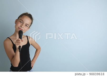 Portrait of a cute teenage girl singing, using microphone. Teenage girl in black t-shirt on an isolated blue background. Karaoke for kids, home entertainment for kids. Singer singing with microphone. Portrait of a cute teenage girl singing, using microphone. Teenage girl in black t-shirt on an isolated blue background. Karaoke for kids, home entertainment for kids. Singer singing with microphone. 98839618
