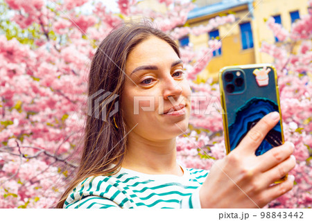 Cheerful young woman talking her picture with her cell phone. Caucasian female model photographing herself with smart phone at spring park. Cheerful young woman talking her picture with her cell phone. Caucasian female model photographing herself with smart phone at spring park. 98843442