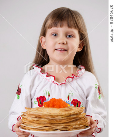A girl in a Ukrainian embroidered dress is holding pancakes with red caviar A girl in a Ukrainian embroidered dress is holding pancakes with red caviar 98845620