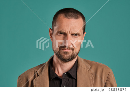 Middle-aged man with beard and mustache, wears black shirt and brown jacket posing against a blue background. Sincere emotions concept. 98853075