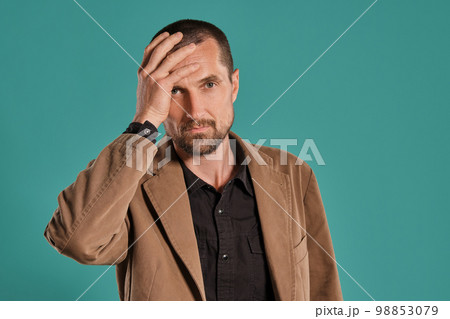 Middle-aged man with beard and mustache, wears black shirt and brown jacket posing against a blue background. Sincere emotions concept. 98853079