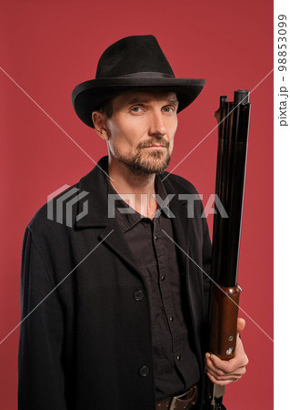 Middle-aged man with beard, mustache, in black jacket and hat, holding a gun while posing against a red background. Sincere emotions concept. Middle-aged man with beard, mustache, in black jacket and hat, holding a gun while posing against a red background. Sincere emotions concept. 98853099