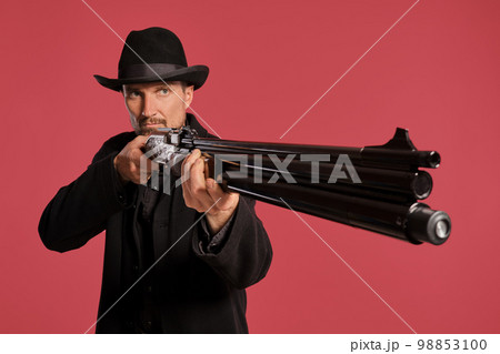 Middle-aged man with beard, mustache, in black jacket and hat, holding a gun while posing against a red background. Sincere emotions concept. 98853100
