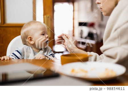 Mother spoon feeding her infant baby boy child sitting in high chair at the dining table in kitchen at home. Mother spoon feeding her infant baby boy child sitting in high chair at the dining table in kitchen at home. 98854611