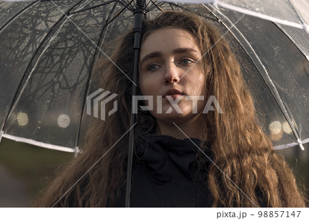 Sadness young woman with lush brown hair under transparent umbrella. Seasonal autumn portrait. Sadness young woman with lush brown hair under transparent umbrella. Seasonal autumn portrait. 98857147