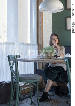 Sad young woman sitting in cafe alone with white cup of tea coffee latte and wait someone. Vertical frame. 98858934