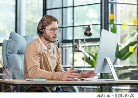 Online psychologist. A young man, a psychologist, a psychotherapist sits in the office in a headset, works at a computer, consults, talks via video call to clients, patients. Online psychologist. A young man, a psychologist, a psychotherapist sits in the office in a headset, works at a computer, consults, talks via video call to clients, patients. 98859720