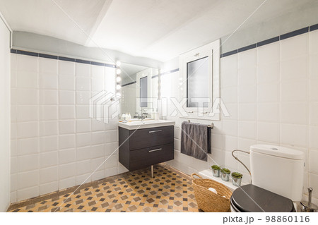 Large bathroom with glossy white walls and beige marble mosaic floor. In the corner is a sink on a vanity with black wooden drawers and a mirror with bright lamps. Near a window for ventilation. Large bathroom with glossy white walls and beige marble mosaic floor. In the corner is a sink on a vanity with black wooden drawers and a mirror with bright lamps. Near a window for ventilation. 98860116