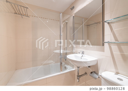Spacious bathroom with soft peach walls, small sink and square mirror. Next to it is white toilet bowl and glass shelves above it for various accessories. On other side is white, clean bathtub. 98860128